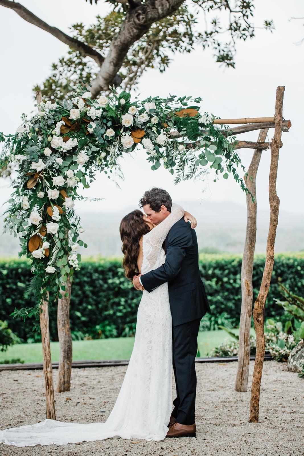 A couple kissing under a timber arbour dressed with white flowers and trailing greenery